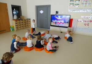 Maluchy siedzą bądź leżą na pomarańczowych poduszkach. Oglądają występ 5-latków na monitorze multimedialnym.